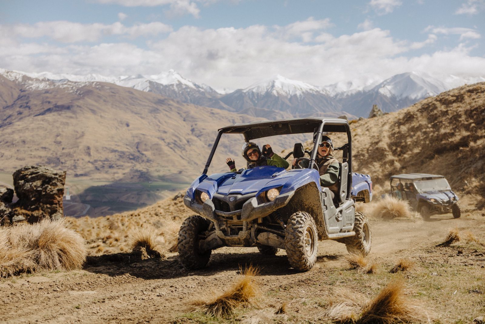 Two riders smiling and waving from a blue Yamaha Viking self-drive off-road buggy on a high-country track in Cardrona Valley, with snow-capped mountains behind